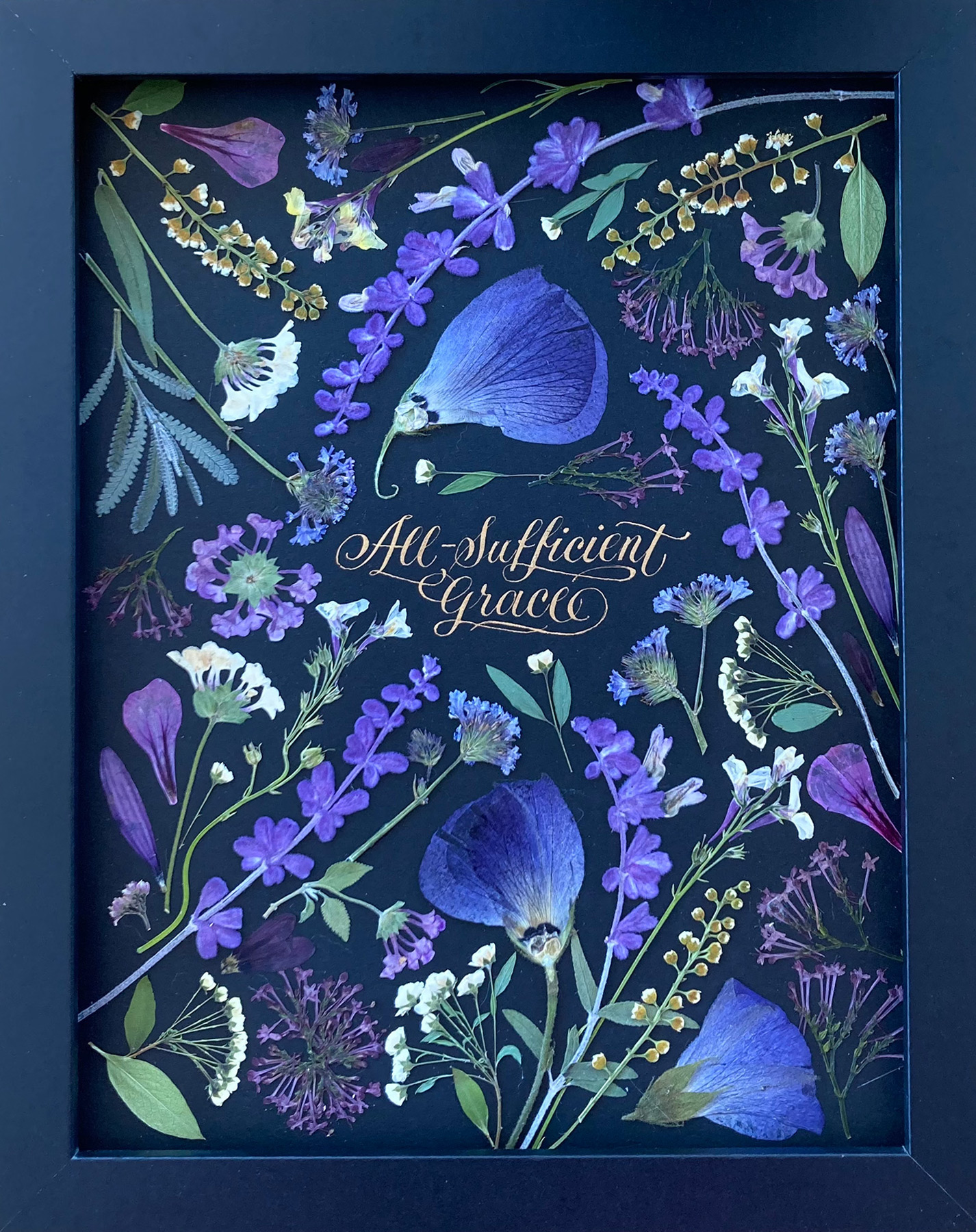 Purple pressed flowers on black paper around gold calligraphy reading All-Sufficient Grace